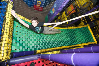 A Boy swinging  on a green net in an indoor play area with colorful padded structures and netting.